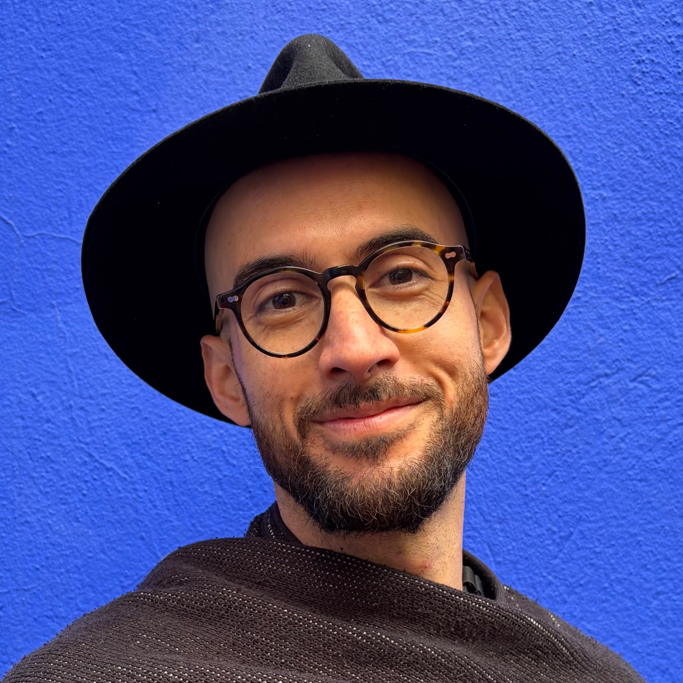 José Rizzo smiling in a black hat and glasses against a vivid blue wall
