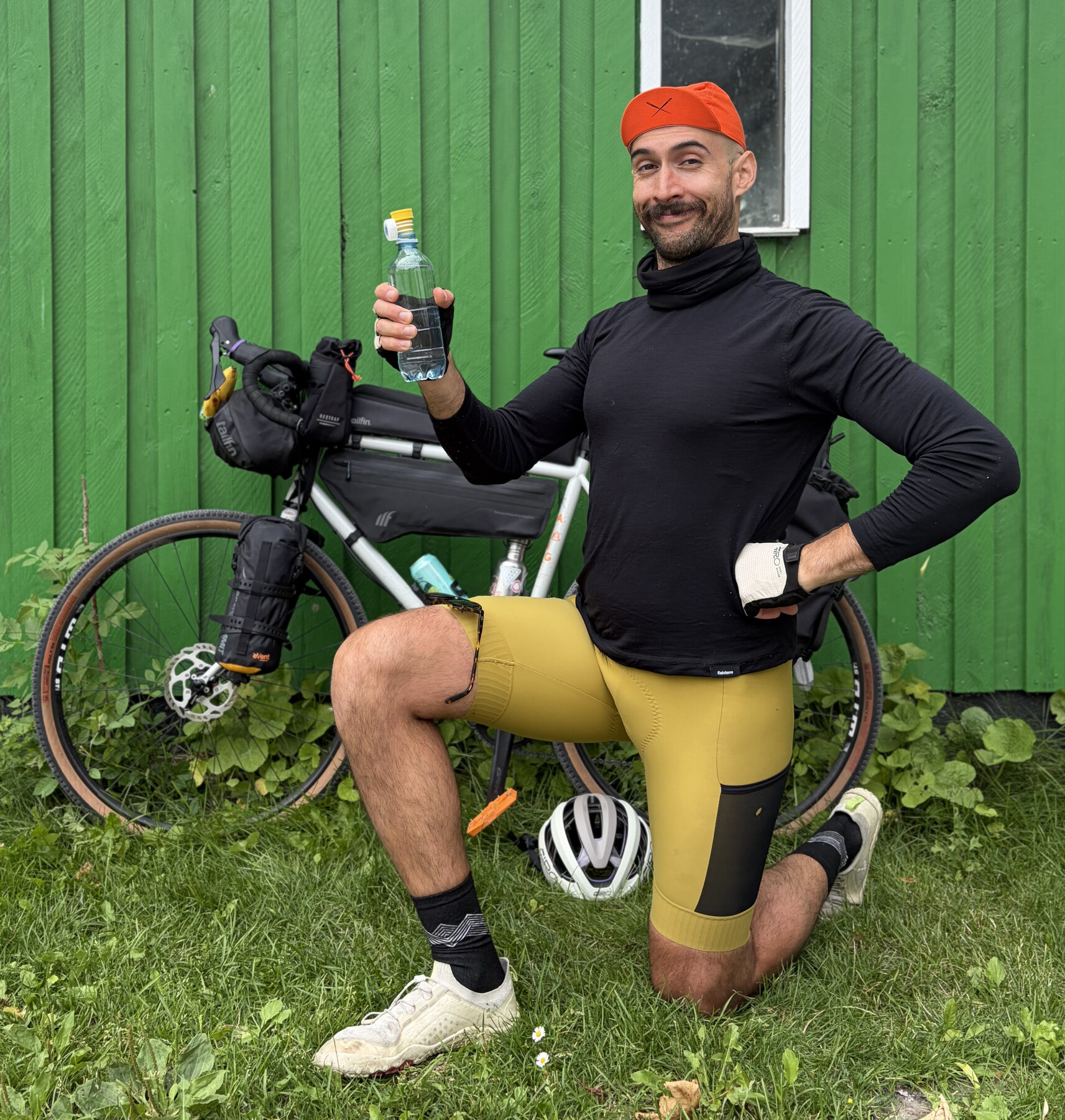 José kneeling beside his loaded bicycle, smiling and raising a water bottle